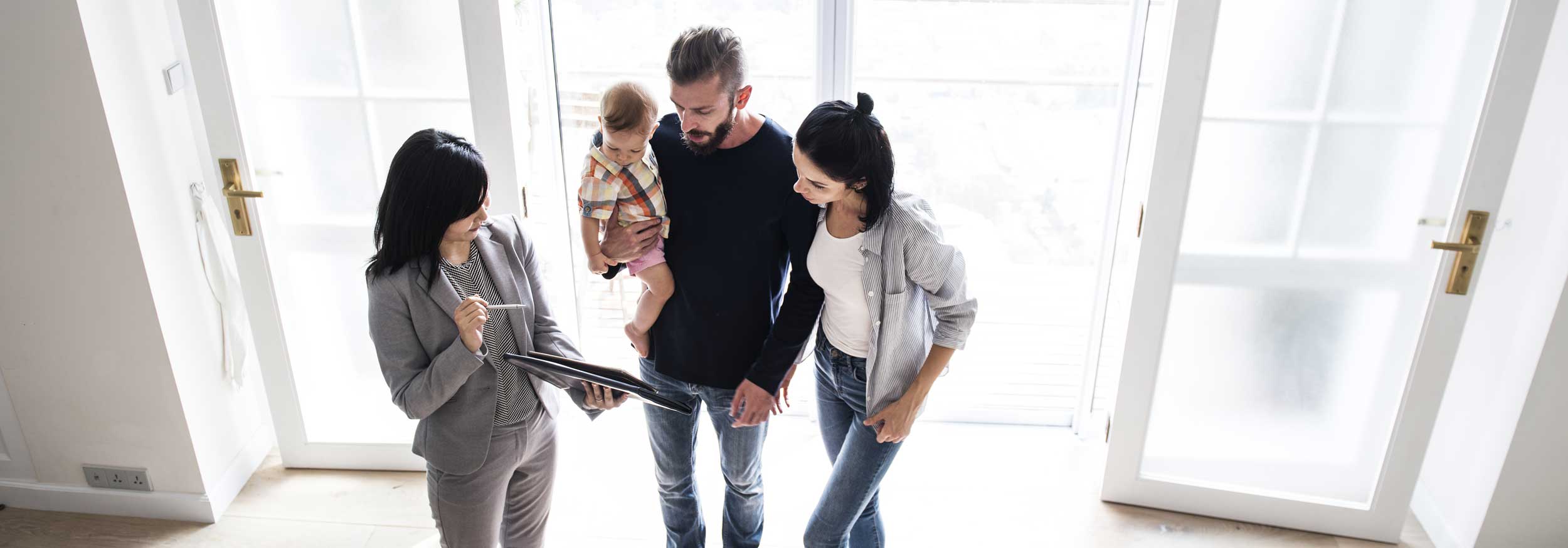A white man and woman holding a baby are talking to a female realtor and reviewing a document in the entrance to a home